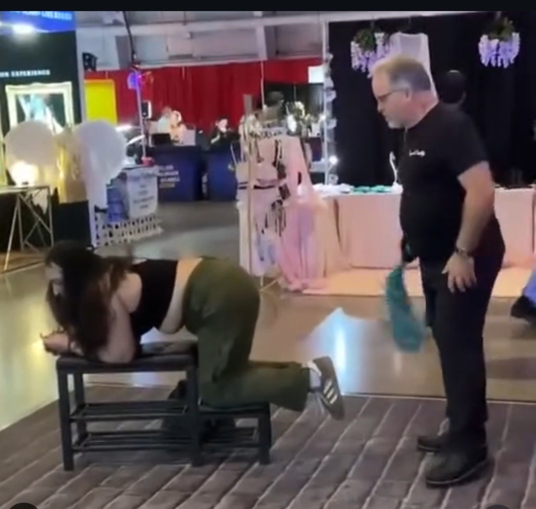 Spank Daddy beside an adult participant at a demonstration bench while holding a teal flogger